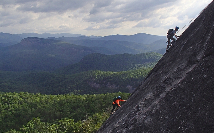 Two people wearing safety gear are secured by ropes as they make their way up a rock incline. Behind them is a vast mountainous area covered in thick green trees. 
